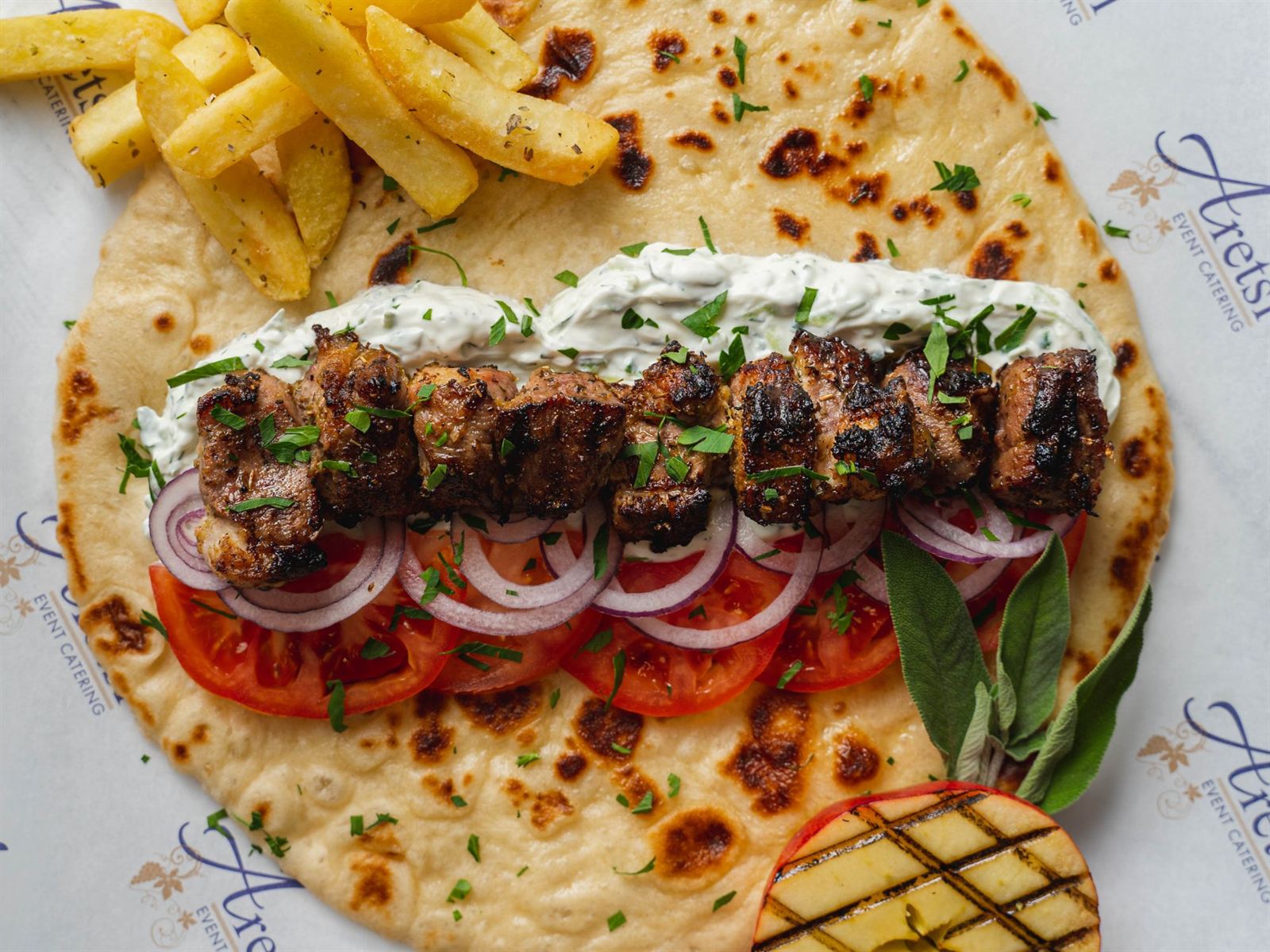 An open Aretsi pork souvlaki pitta with tzatziki, tomato, red onion, fries and a grilled peach on Aretsi paper