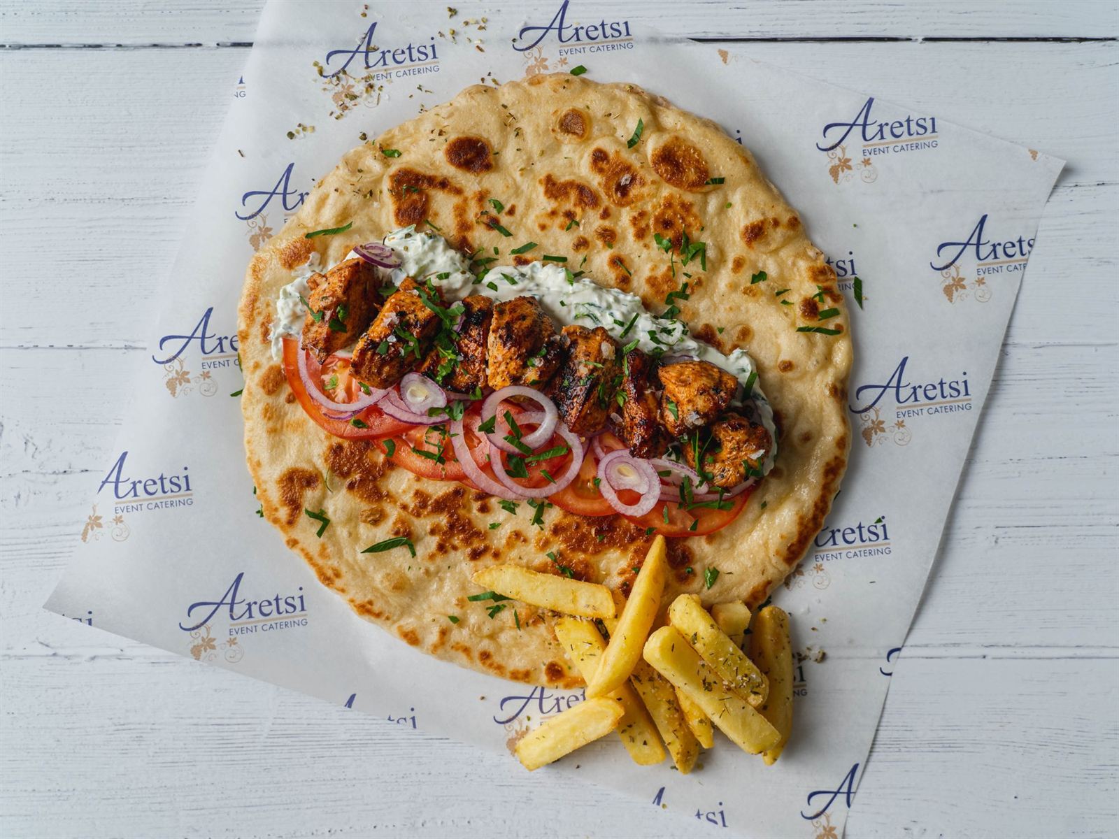 An open Aretsi chicken souvlaki pitta with tzatziki, tomato, red onion and oregano fries on Aretsi paper
